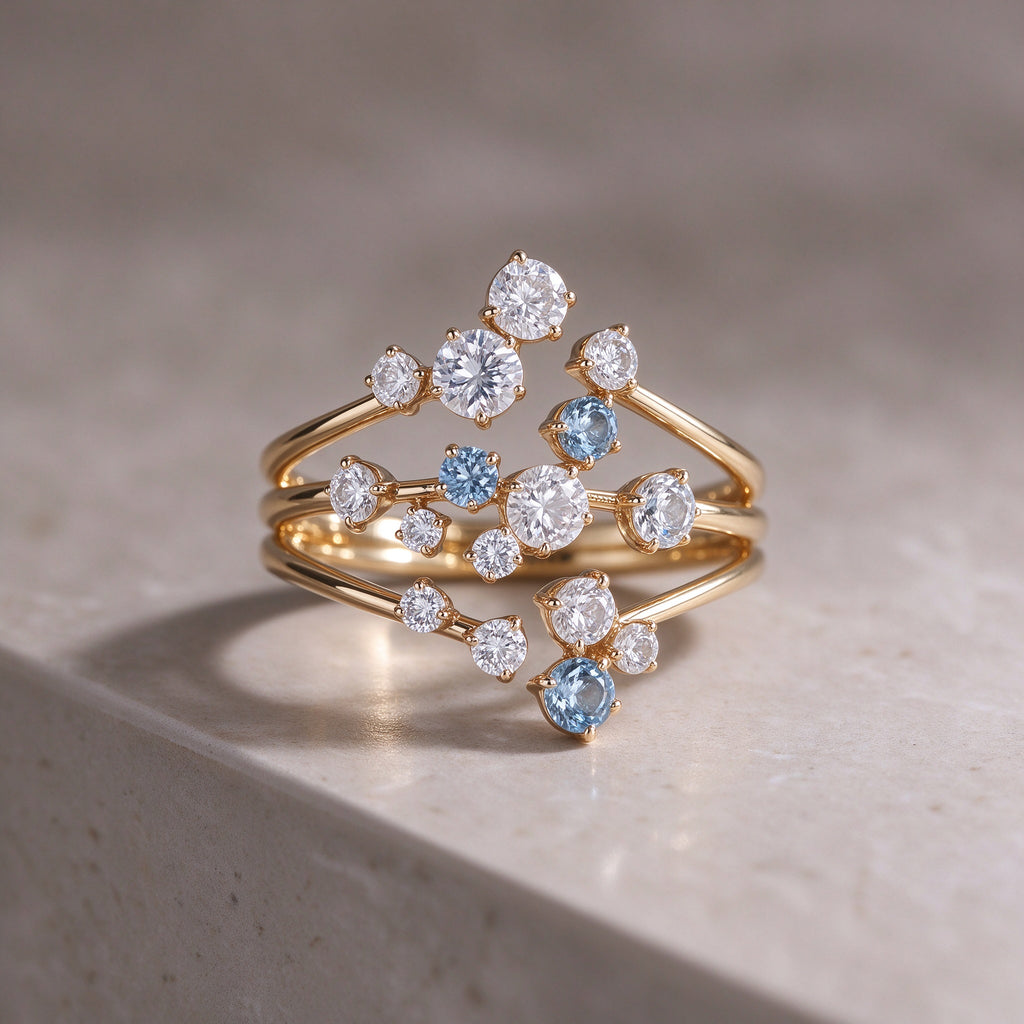The Starpath Ring in 18K gold vermeil over sterling silver, with white moissanite and pale lab-created blue sapphires set along three sweeping gold arcs, resting on a light stone surface.