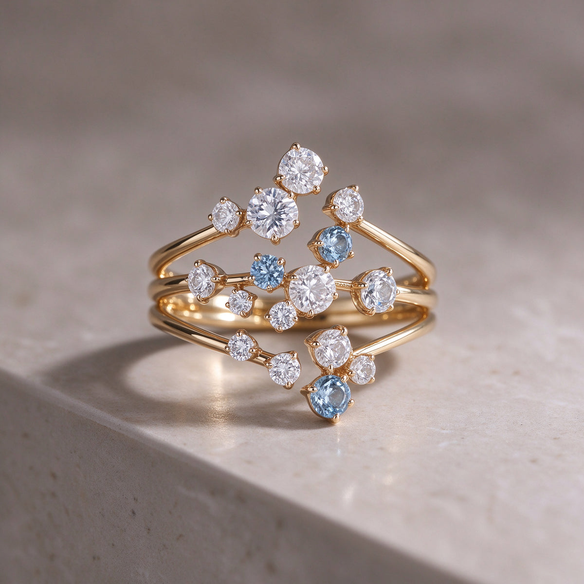 The Starpath Ring in 18K gold vermeil over sterling silver, with white moissanite and pale lab-created blue sapphires set along three sweeping gold arcs, resting on a light stone surface.
