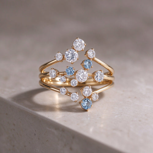 The Starpath Ring in 18K gold vermeil over sterling silver, with white moissanite and pale lab-created blue sapphires set along three sweeping gold arcs, resting on a light stone surface.